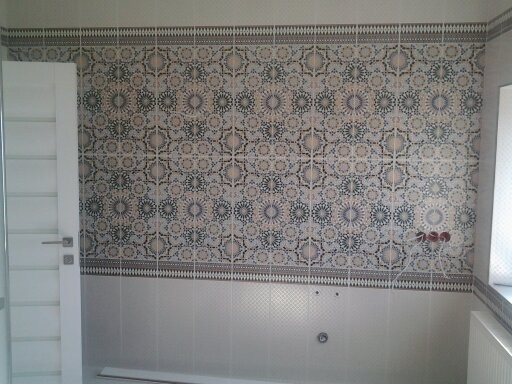 Ściana w łazience wyłożona dekoracyjnymi płytkami ceramicznymi w odcieniach beżu, szarości i granatu, z widoczną białą ramą drzwi po lewej stronie i wystającymi rurami instalacji wodnej na środku...