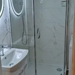 Nowoczesna łazienka po remoncie: kabina prysznicowa narożna ze szklanymi drzwiami, umywalka z szafką, lustro z podświetleniem LED i marmurowe płytki na ścianach.