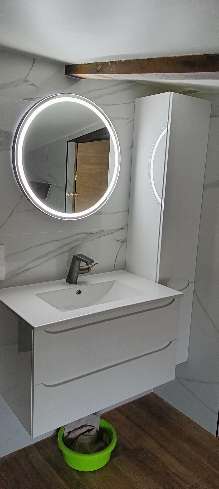 Nowoczesna łazienka z okrągłym lustrem LED, białą szafką pod umywalkę z szufladami i wysoką szafką boczną, wykończona płytkami imitującymi marmur, z widocznym fragmentem drewnianej belki stropowej