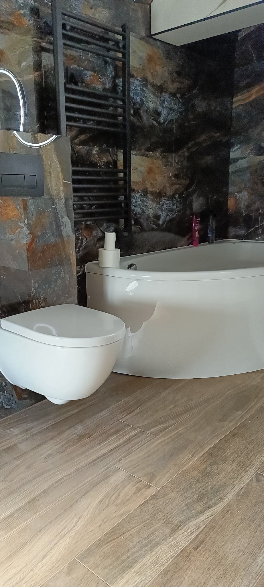 Nowoczesna, biała toaleta podwieszana i wanna z hydromasażem na tle marmurowej ściany i drewnopodobnej podłogi w łazience.