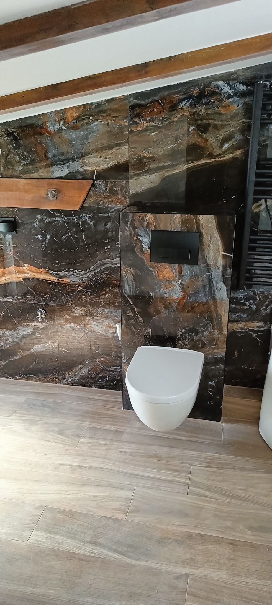 Nowoczesna łazienka z podwieszaną toaletą, ściany i podłoga wyłożone płytkami imitującymi marmur w odcieniach brązu i szarości, drewniane elementy dekoracyjne.