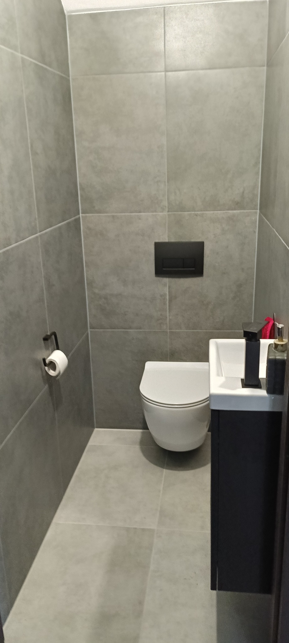 Nowoczesna toaleta w szarościach: wisząca muszla, minimalistyczna umywalka z czarną armaturą, płytki imitujące kamień, uchwyt na papier toaletowy.