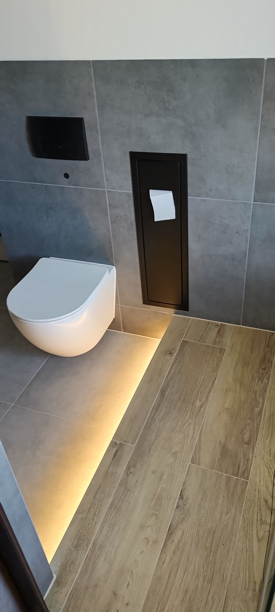 Nowoczesna łazienka z podświetlaną podłogą i podwieszaną toaletą. Szare płytki na ścianach, drewnopodobne na podłodze. Minimalistyczny design.