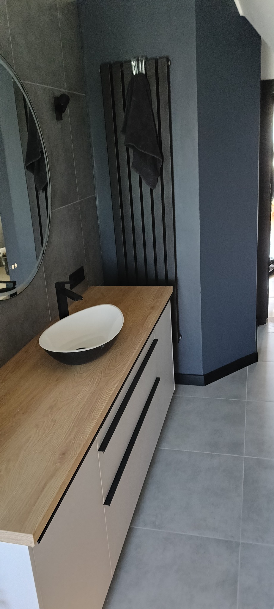 Nowoczesna łazienka z umywalką nablatową na szafce z drewnianym blatem, czarną armaturą i grzejnikiem drabinkowym. Szare płytki i minimalistyczny design.