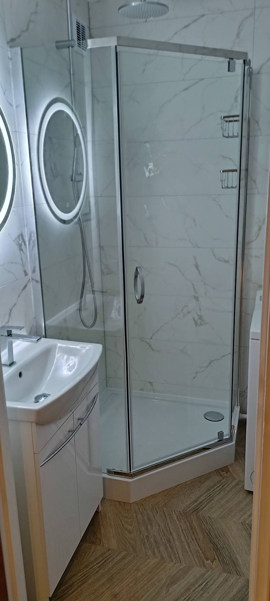 Nowoczesna łazienka po remoncie: kabina prysznicowa narożna ze szklanymi drzwiami, umywalka z szafką, lustro z podświetleniem LED i marmurowe płytki na ścianach.