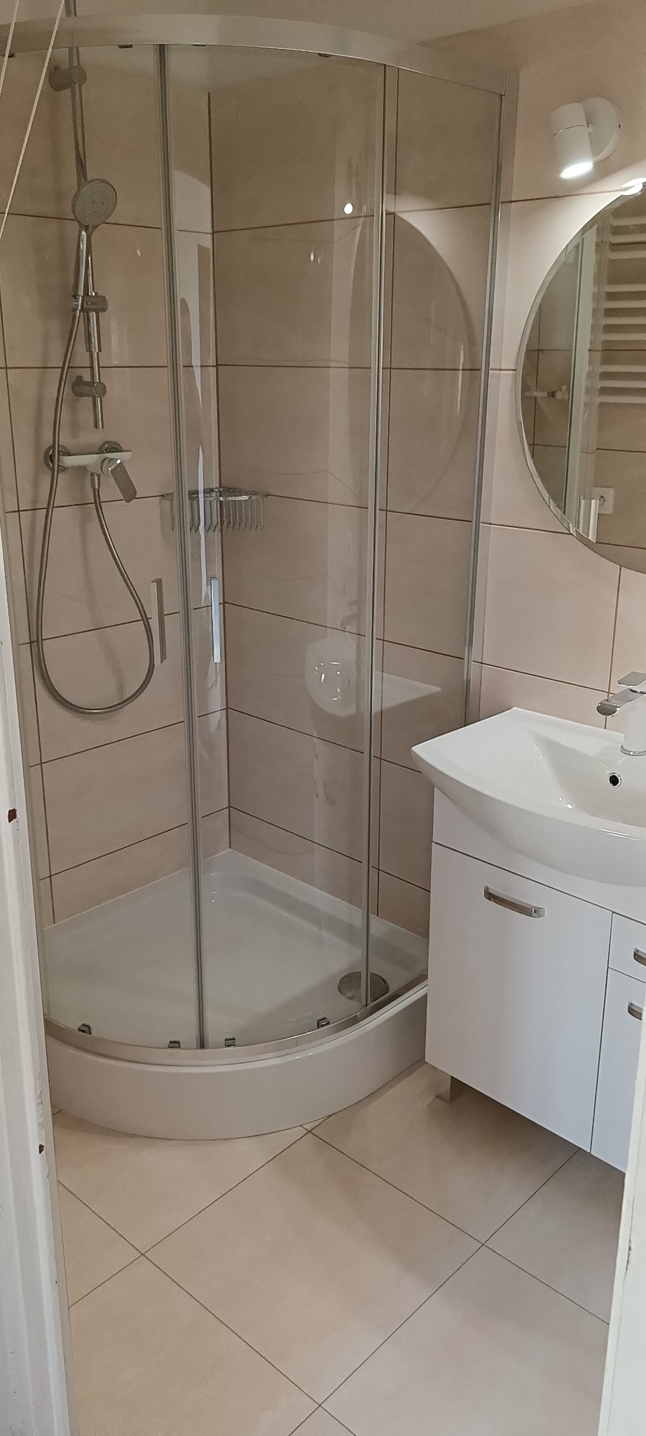 Jasna łazienka po remoncie: narożna kabina prysznicowa z chromowanym zestawem, umywalka z szafką i owalne lustro z oświetleniem. Beżowe płytki na ścianach i podłodze.