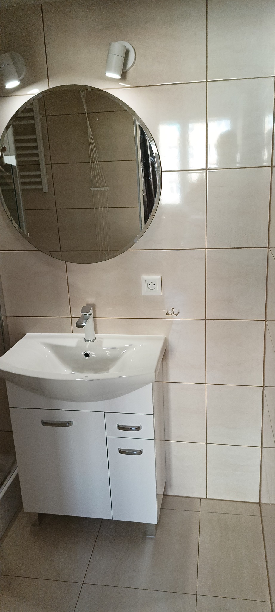 Nowoczesna umywalka z szafką w jasnej łazience, okrągłe lustro z podświetleniem i beżowe płytki tworzą minimalistyczny wystrój.