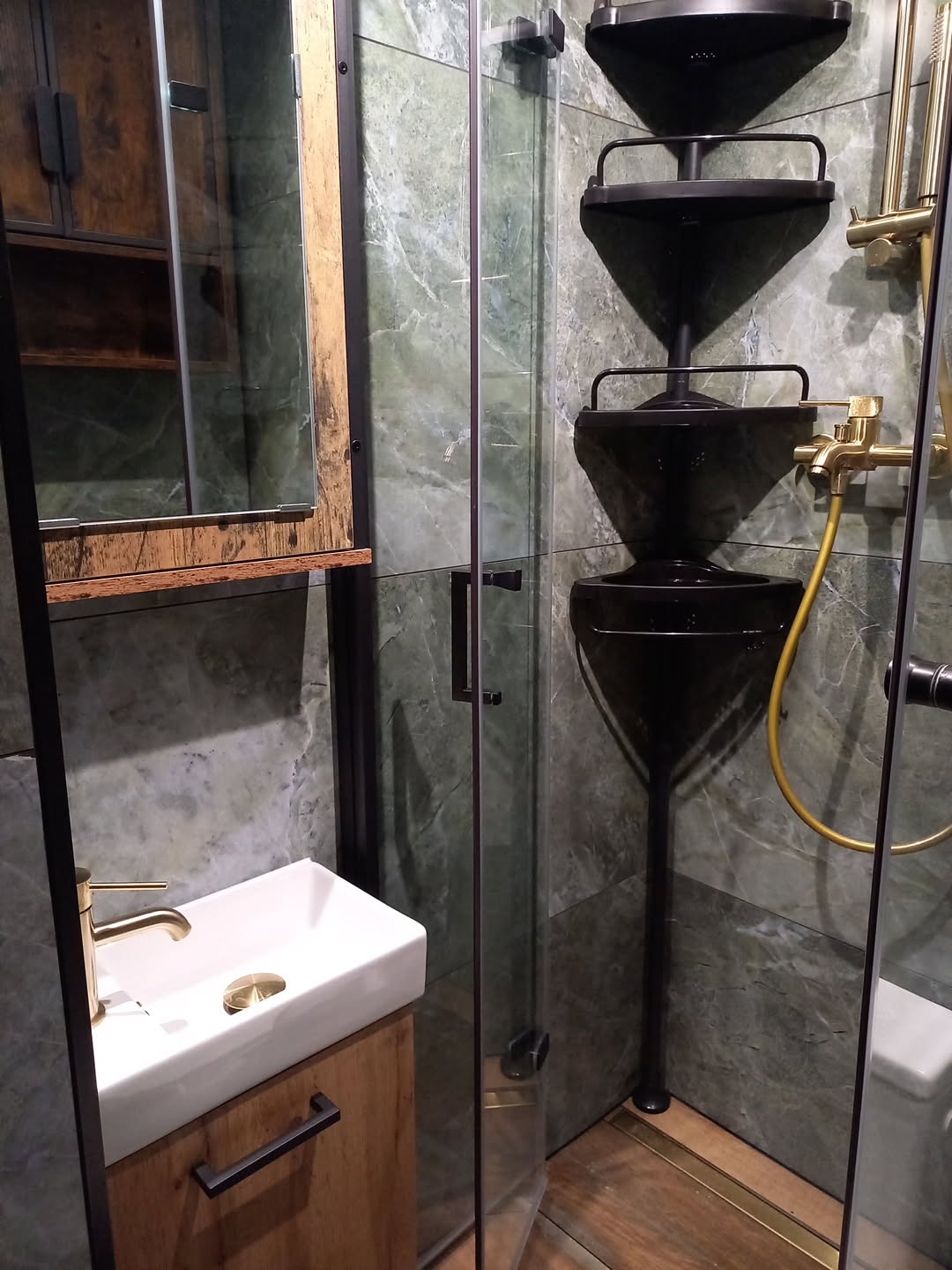 Nowoczesna łazienka z kabiną prysznicową, umywalką na drewnianej szafce i zielonymi marmurowymi płytkami. Czarna armatura i półki narożne dodają elegancji.