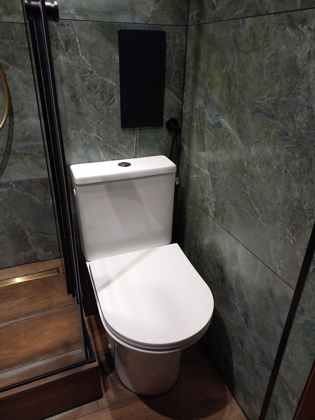 Nowoczesna toaleta w łazience wykończonej marmurowymi płytkami w odcieniach zieleni, z czarnymi akcentami armatury i zabudowy. Widoczny fragment kabiny prysznicowej.