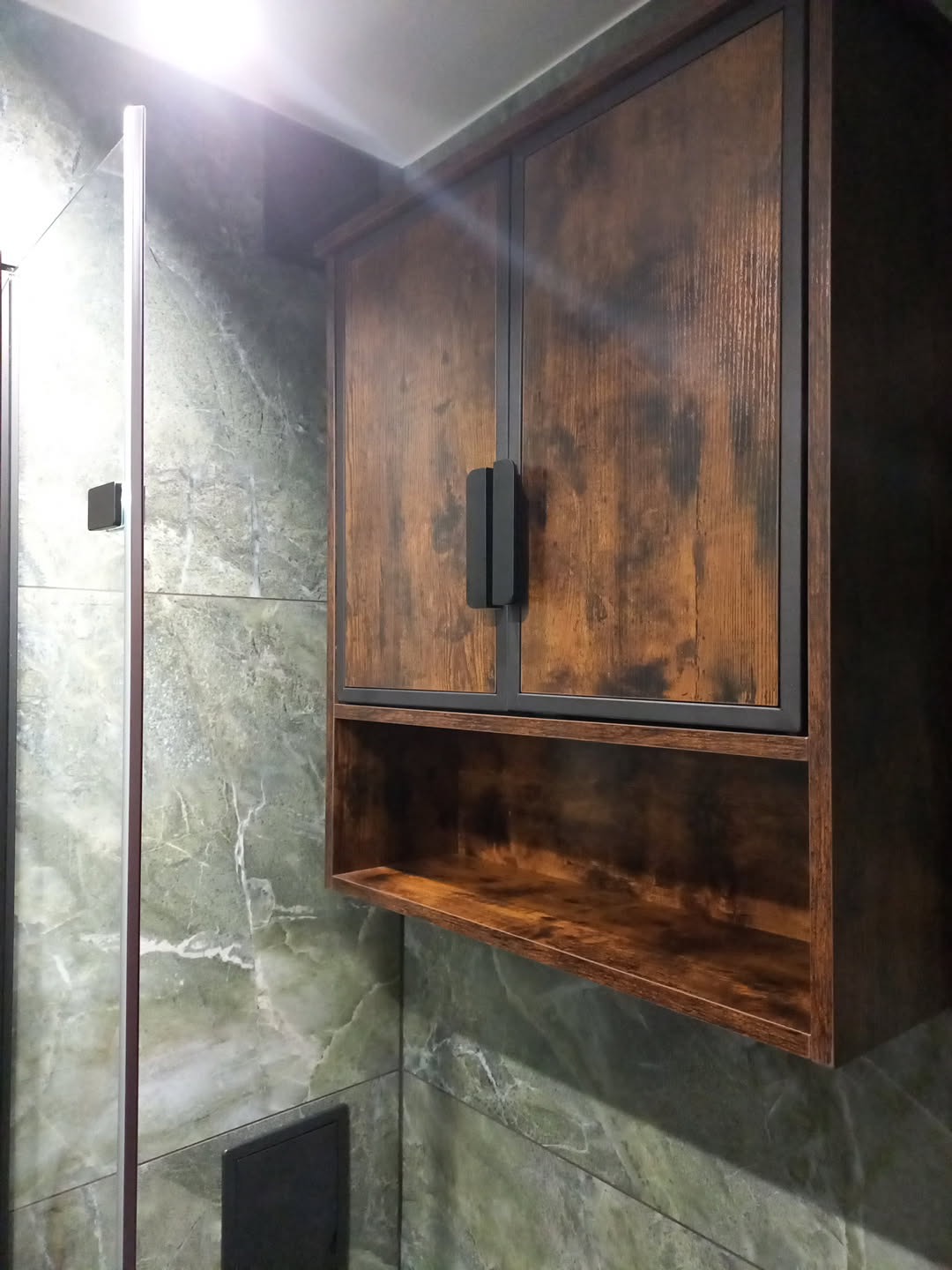 Rustykalna szafka wisząca z półką w łazience z marmurowymi płytkami. Czarny, podwójny uchwyt. Widoczny fragment kabiny prysznicowej.