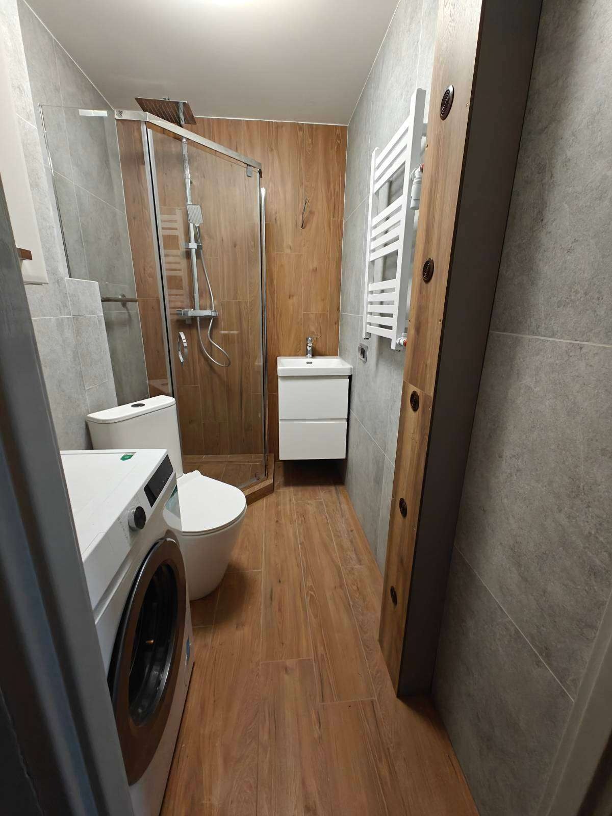 Nowoczesna łazienka po remoncie: kabina prysznicowa, umywalka z szafką, grzejnik drabinkowy, pralka. Płytki imitujące drewno i szarość. Ujęcie z perspektywy wejścia.