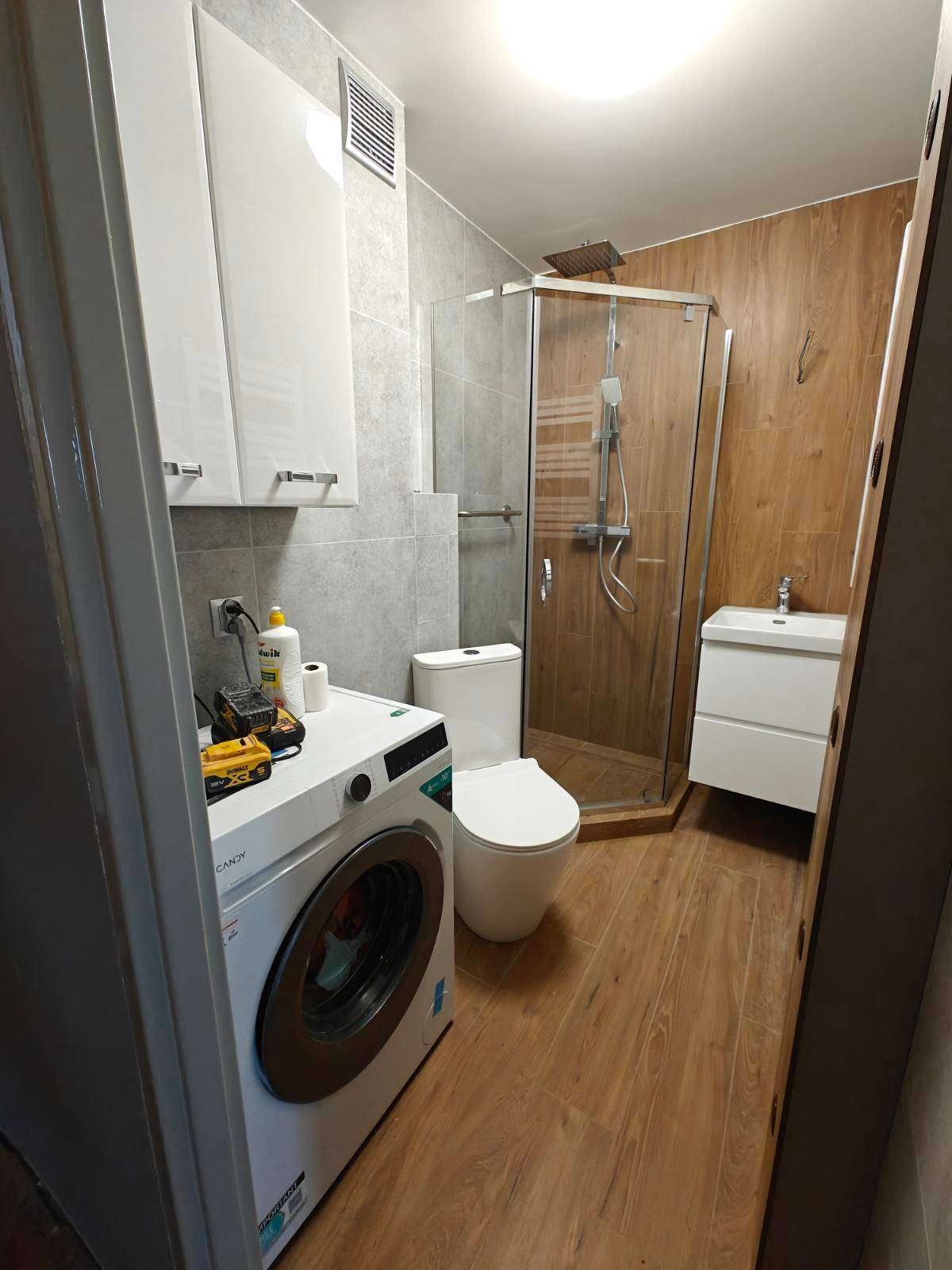Wykończona łazienka z kabiną prysznicową, umywalką, toaletą i pralką. Na pralce leżą narzędzia. Płytki imitujące drewno na podłodze i ścianie kabiny.