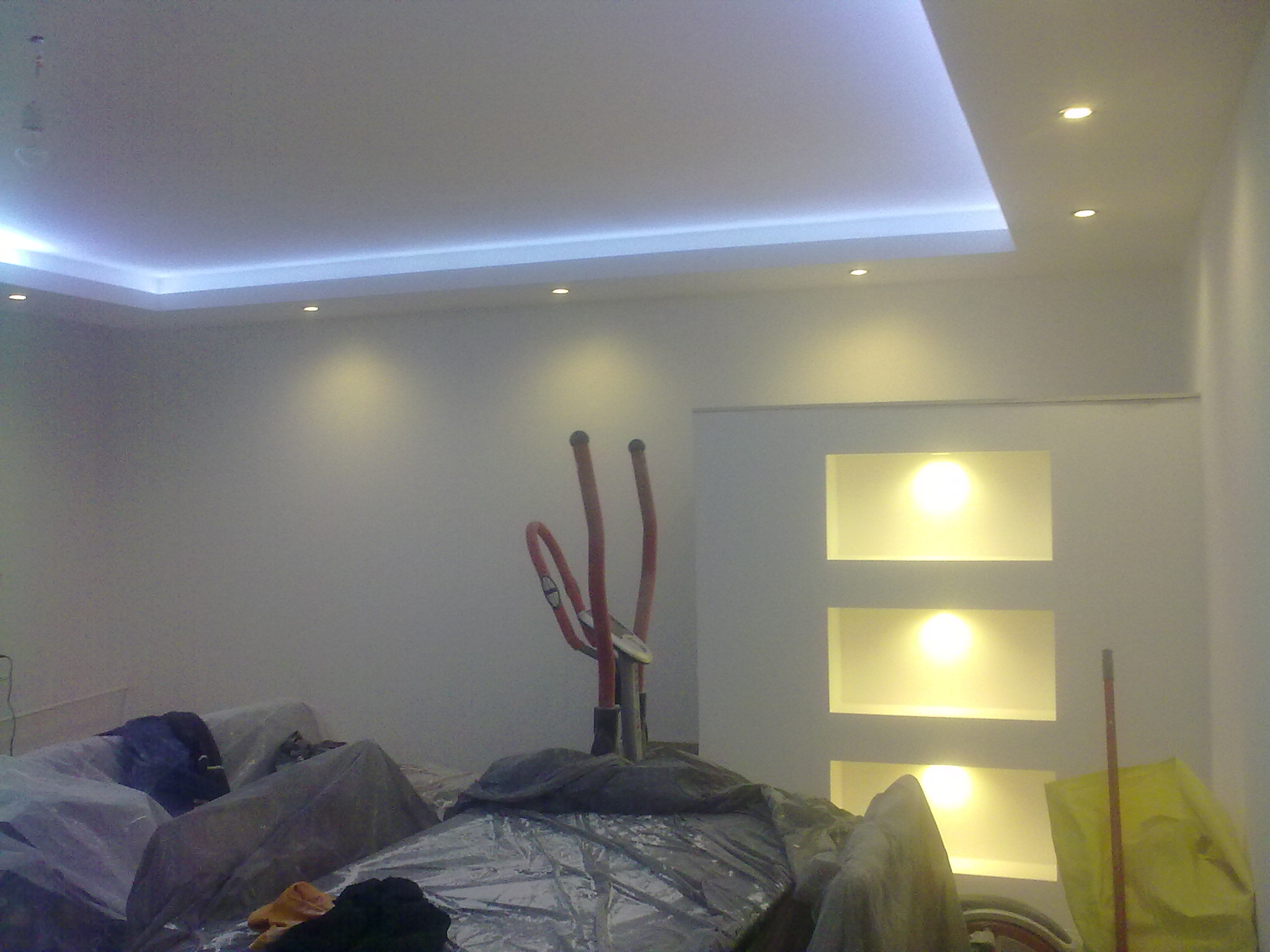 Wykończony sufit podwieszany z płyt karton gips z oświetleniem LED i punktowym, widoczne przygotowania do dalszych prac wykończeniowych, zabezpieczone folią meble.