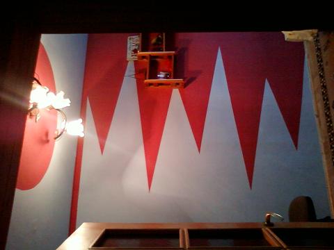 Wnętrze pokoju z dekoracyjnym malowaniem ściany w czerwono-białe trójkąty, oświetlone kinkietem w kształcie kwiatów i z drewnianą półką na ścianie.