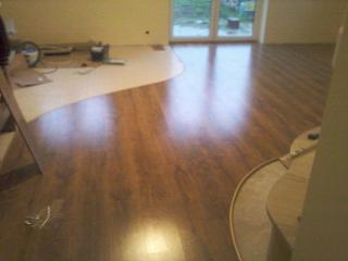 Wnętrze pokoju w trakcie montażu brązowych paneli podłogowych, widoczne narzędzia i częściowo ułożona podłoga z charakterystycznym, zakrzywionym przejściem do przygotowanej powierzchni.