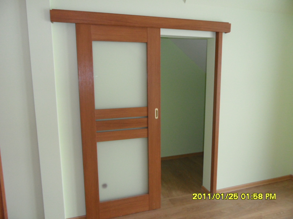 Drewniane drzwi przesuwne z matowym szkłem, zamontowane w jasnym pomieszczeniu z widocznym wejściem do kolejnego pokoju z zielonymi ścianami i podłogą z paneli.