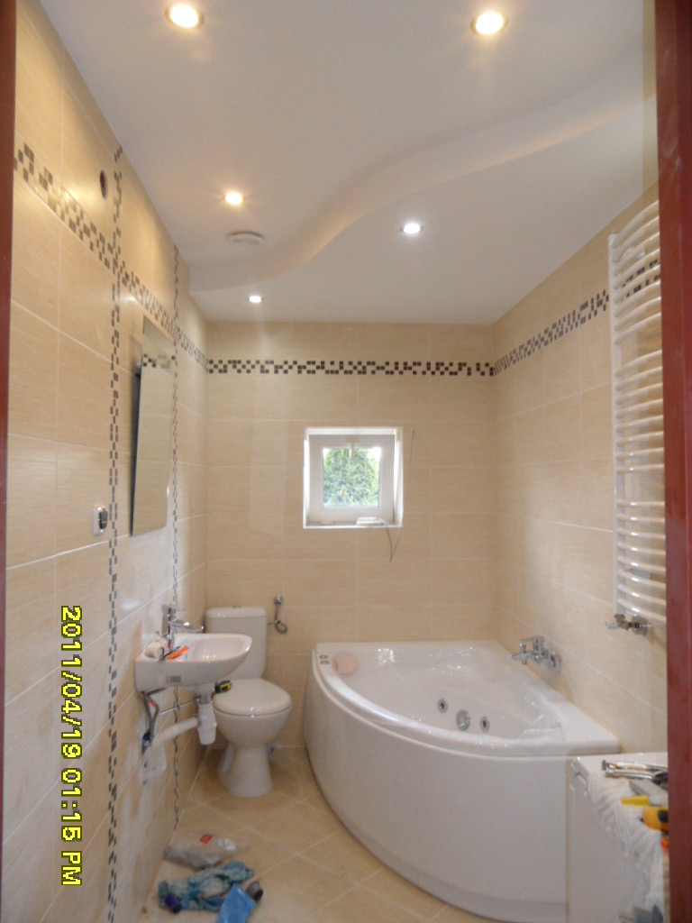 Jasna łazienka po remoncie z narożną wanną z hydromasażem, beżowymi płytkami z dekoracyjnym paskiem i podwieszanym sufitem z oświetleniem punktowym.