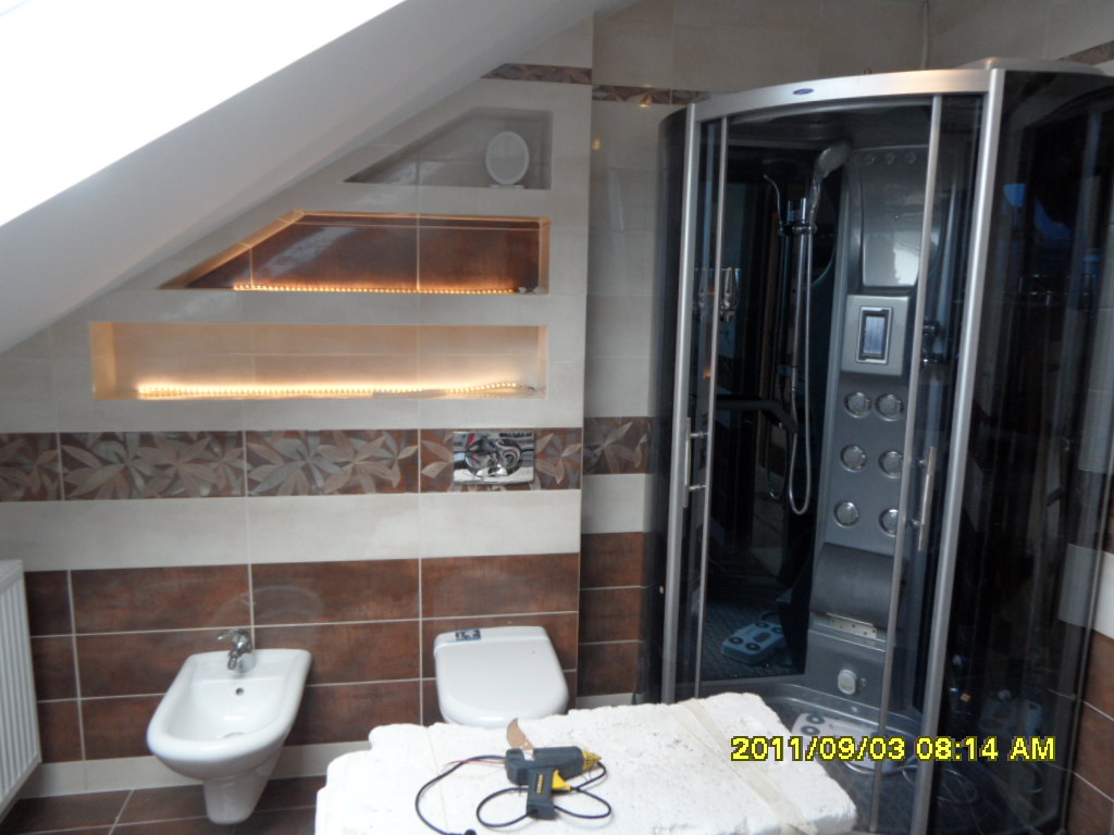 Łazienka na poddaszu z kabiną prysznicową, bidetem i toaletą; w tle widoczne podświetlane wnęki w ścianie, na pierwszym planie styropian i pistolet do kleju.