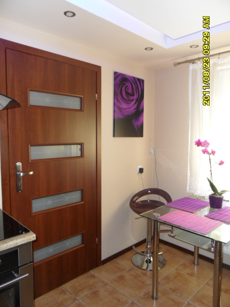 Wnętrze kuchni z brązowymi płytkami podłogowymi, drewnianymi drzwiami z przeszkleniami i szklanym stołem barowym z fioletowymi podkładkami. Na ścianie wisi obraz z motywem fioletowej róży.
