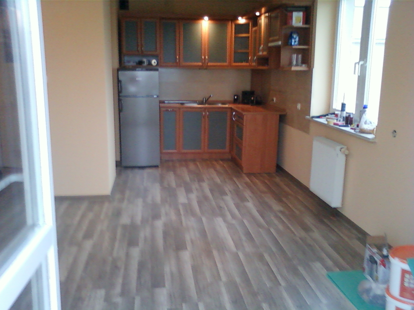 Kuchenne panele podłogowe w odcieniach szarości z widoczną strukturą drewna, świeżo po montażu, w tle zabudowa kuchenna w odcieniach brązu i beżu, częściowo widoczny grzejnik i okno.