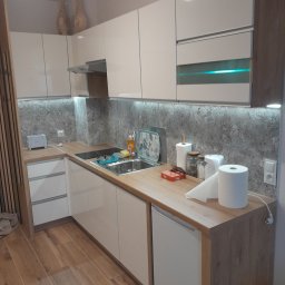 OLMIR Mirosław Sochacki - Jasna kuchnia z drewnianymi blatami i szarym backsplashiem, oświetlenie LED pod szafkami, drewniana podłoga, widoczne akcesoria kuchenne i dekoracyjne wazony na szafkach.