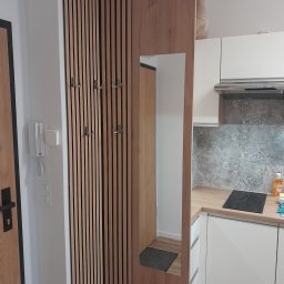 OLMIR Mirosław Sochacki - Nowoczesne przedpokoju z drewnianą ścianką działową z haczykami, lustrem wbudowanym w panel i widokiem na kuchnię z białą zabudową i drewnianym blatem.