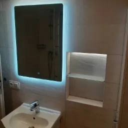 Nowoczesna łazienka z umywalką, podświetlanym lustrem LED i wnęką z półkami, wykończona płytkami imitującymi kamień.