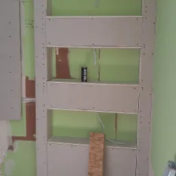 Konstrukcja z płyty gipsowo-kartonowej tworząca półki w trakcie montażu, widoczne przewody elektryczne, puszka napoju energetycznego na jednej z półek, fragment drabiny i deski na podłodze.