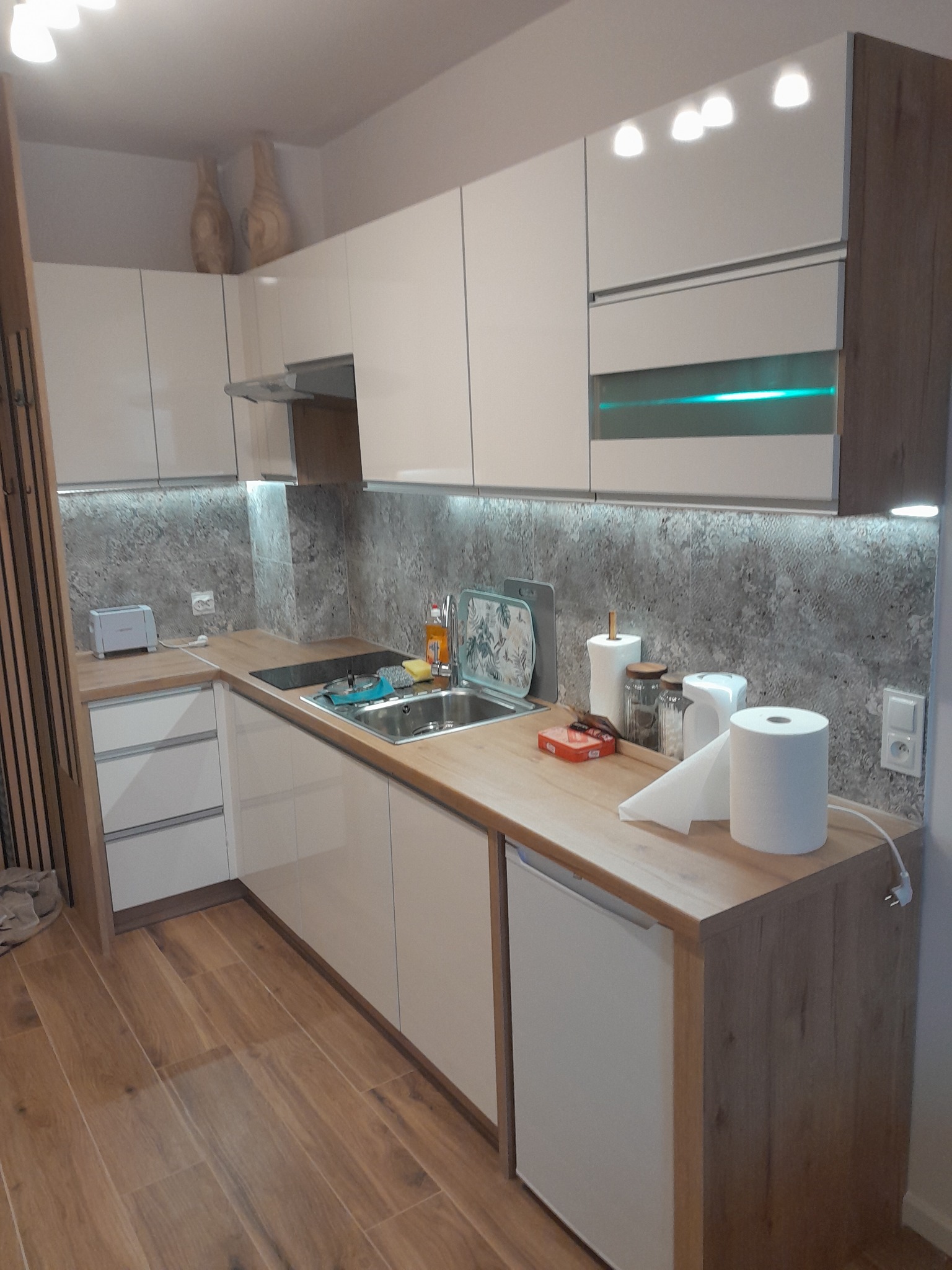 Jasna kuchnia z drewnianymi blatami i szarym backsplashiem, oświetlenie LED pod szafkami, drewniana podłoga, widoczne akcesoria kuchenne i dekoracyjne wazony na szafkach.