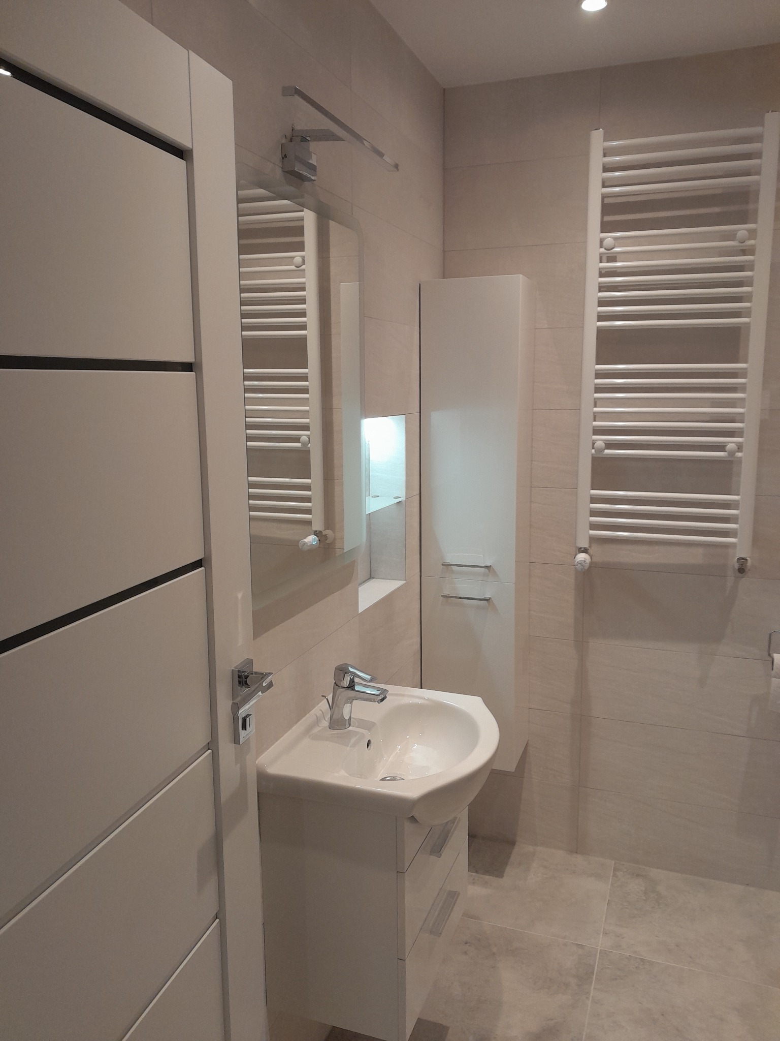 Jasna, minimalistyczna łazienka z umywalką na szafce, grzejnikiem drabinkowym i lustrem, wykończona beżowymi płytkami.