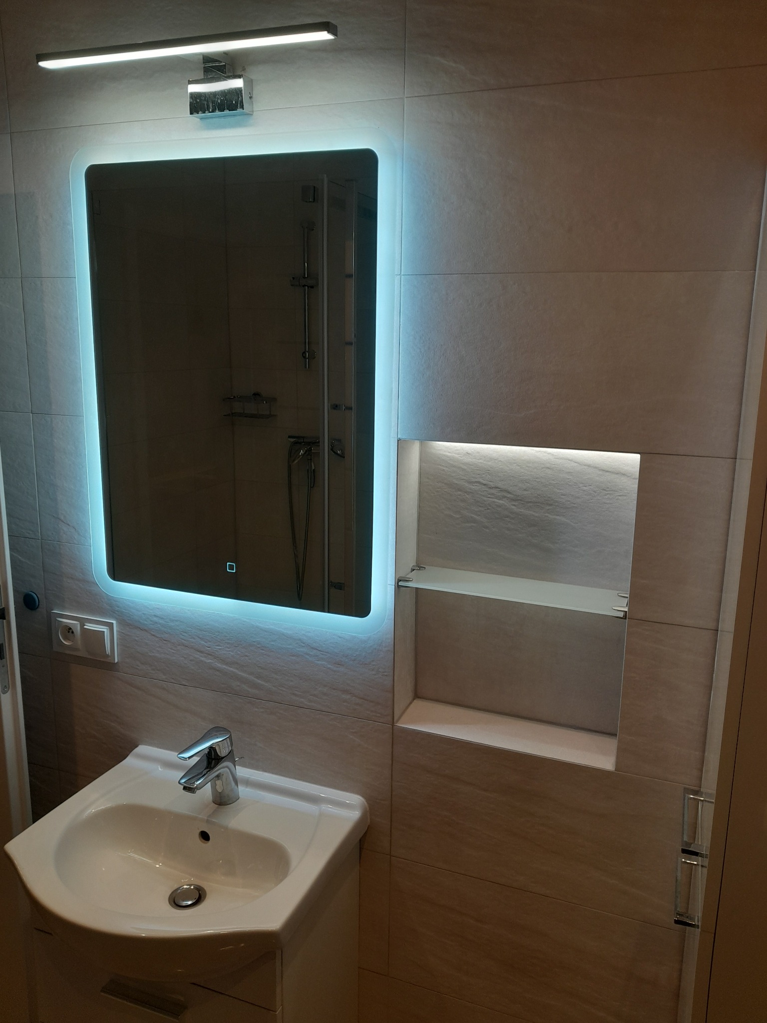 Nowoczesna łazienka z umywalką, podświetlanym lustrem LED i wnęką z półkami, wykończona płytkami imitującymi kamień.