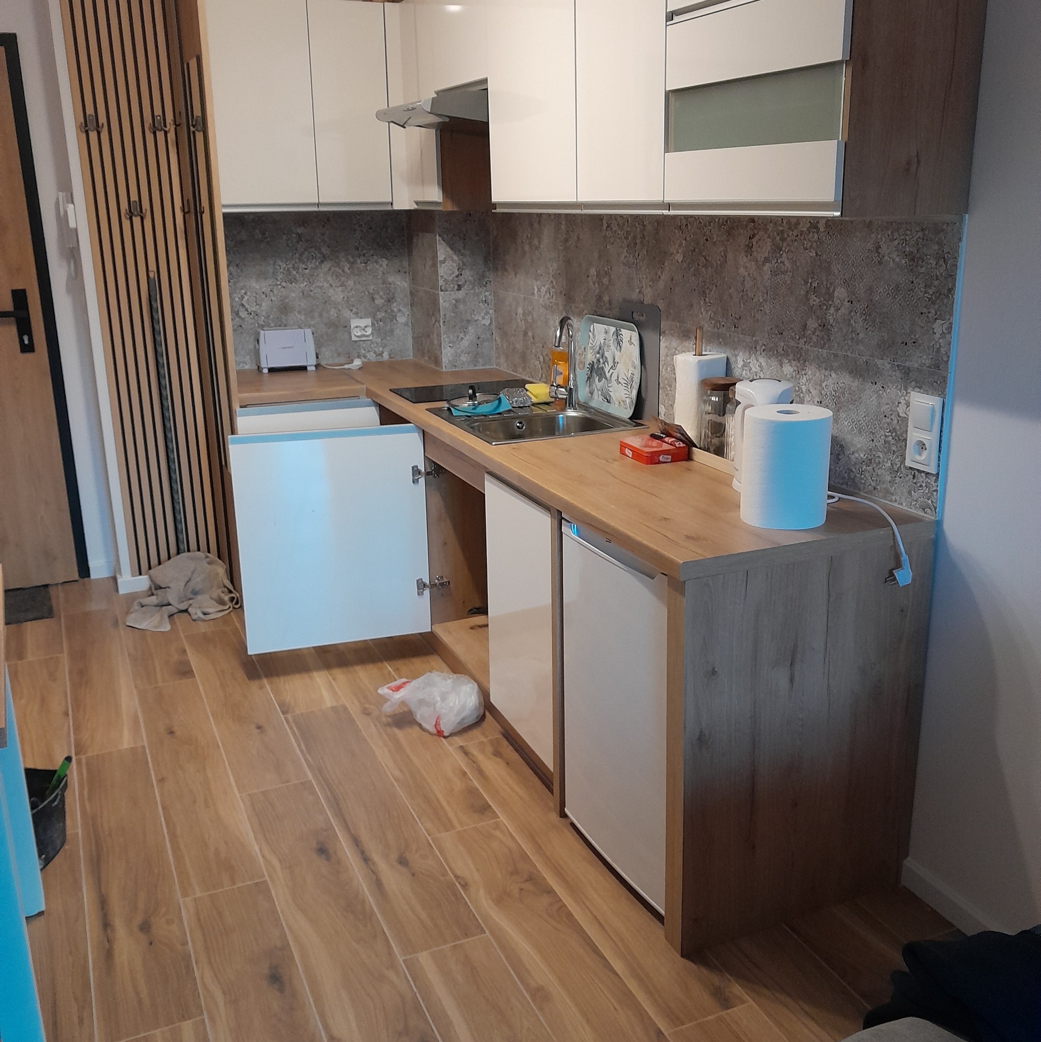 Wykończona kuchnia w Jeleniej Górze z drewnianą podłogą, jasnymi szafkami i szarym blatem. Widoczny otwarty front szafki, zlew ze środkami czystości i akcesoriami oraz reling z wieszakami na ścianie.