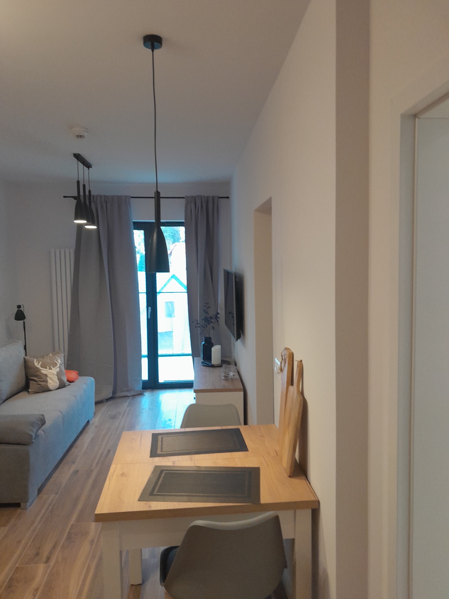 Jasne wnętrze mieszkania z drewnianą podłogą, stołem jadalnym z szarymi podkładkami, szarą sofą z poduszką z motywem jelenia, oknem z szarymi zasłonami i nowoczesnym oświetleniem wiszącym.