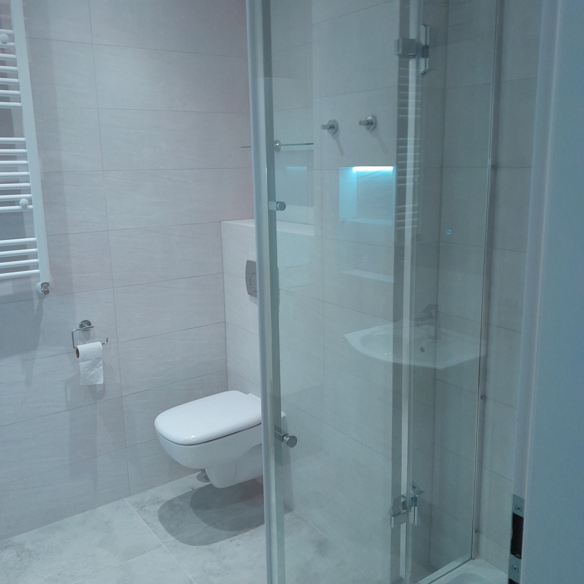 Nowoczesna łazienka z kabiną prysznicową ze szkła hartowanego, podwieszaną toaletą i grzejnikiem drabinkowym w kolorze białym, wykończona jasnymi płytkami ceramicznymi.