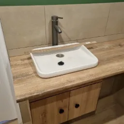 Biała, prostokątna umywalka nablatowa z szarą baterią na blacie imitującym drewno, zamontowana na szafce z dwojgiem drzwi o fakturze drewna, widoczna listwa przypodłogowa i płytki na ścianie.