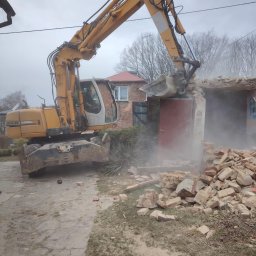RAFSED - Żółta koparka Liebherr w trakcie rozbiórki murowanego budynku. Widoczne gruzy i pył. W tle budynek mieszkalny z czerwonym dachem i drzewa bez liści.