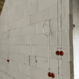 Surowa ściana z betonu komórkowego z wyfrezowanymi bruzdami i poprowadzonymi przewodami elektrycznymi oraz zamontowanymi puszkami pod gniazdka, widoczne oznaczenia ołówkiem na ścianie.