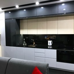 Vaker Paweł Głownicki - Nowoczesna kuchnia z białymi, szarymi i czarnymi frontami szafek, czarnym blatem, szklanym panelem nad blatem z widokiem miasta i oświetleniem punktowym, widziana z perspektywy salonu.