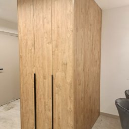 Vaker Paweł Głownicki - Wysoka szafa z drewnopodobnymi frontami i minimalistycznymi czarnymi uchwytami, przylegająca do ściany, widoczna część marmurowej podłogi i krzesła w tle.