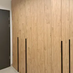 Wysoka szafa wnękowa z jasnego drewna z ciemnymi, pionowymi uchwytami, ustawiona w rogu pomieszczenia z marmurową podłogą i szarą ścianą z drzwiami.