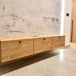 Vaker Paweł Głownicki - Wisząca szafka RTV z drewnianymi frontami na tle ściany o surowym wykończeniu, widoczne czarne uchwyty i fragment szafy wnękowej z identycznego materiału.