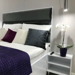 Nowoczesna sypialnia w Mysłowicach z tapicerowanym zagłówkiem w kolorze grafitowym, białą pościelą, dekoracyjnymi poduszkami w odcieniach fioletu i szarości oraz białą szafką nocną z orchideą...