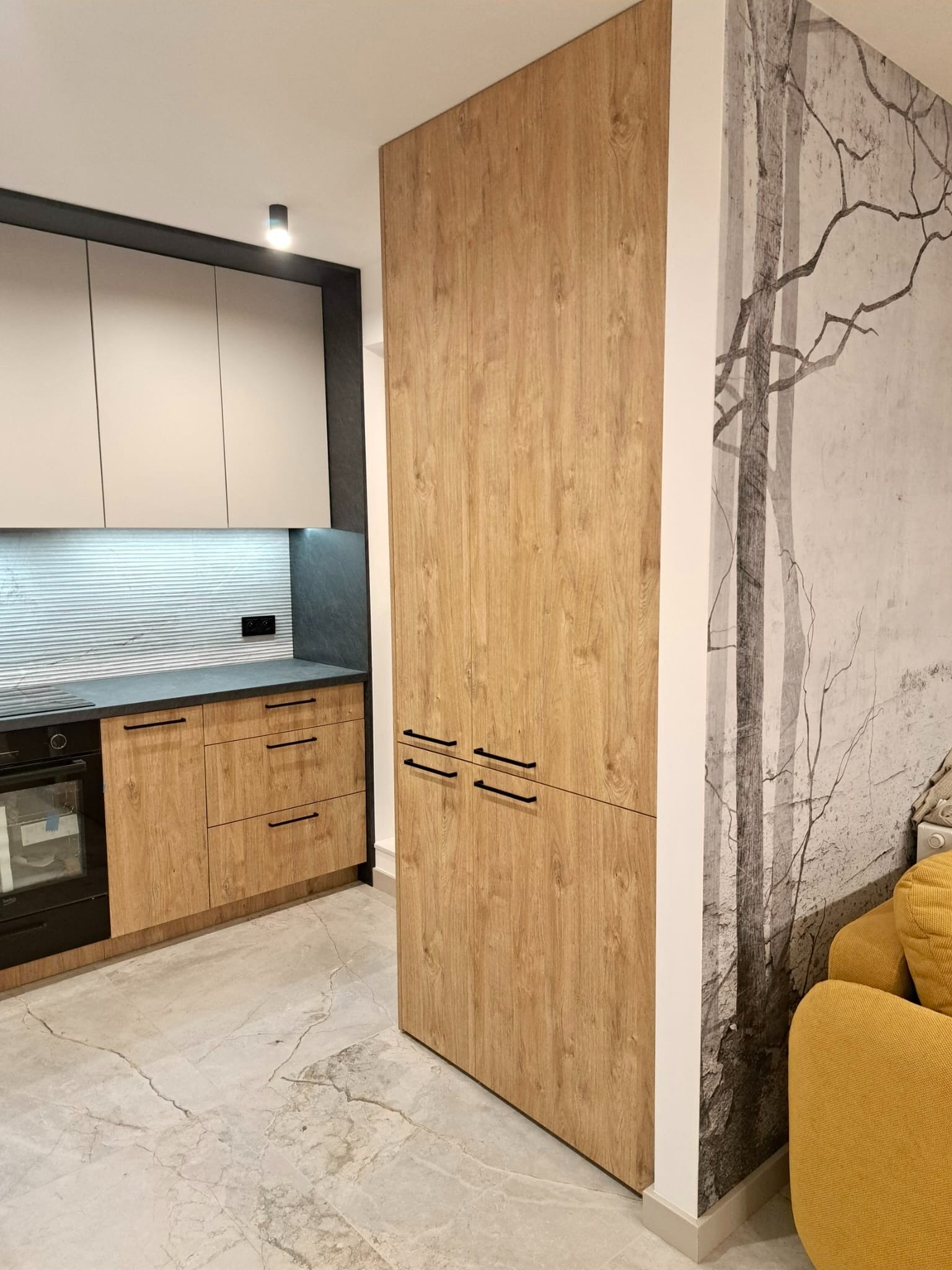 Nowoczesna kuchnia z frontami w kolorze jasnego drewna i szarymi górnymi szafkami, czarny blat i sprzęt AGD, wysoka zabudowa z drewnianymi frontami, na ścianie tapeta z motywem drzew, marmurowa...