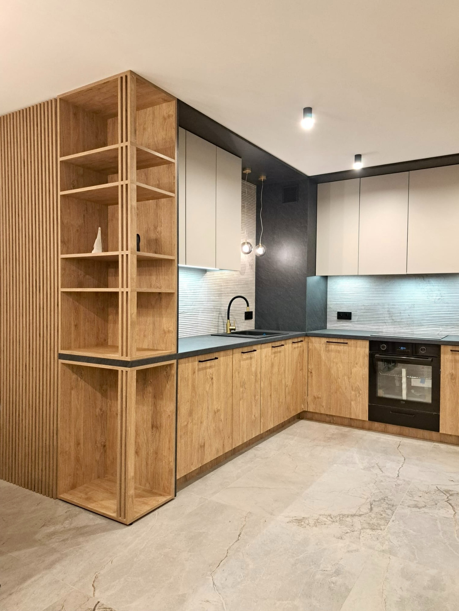 Nowoczesna kuchnia z drewnianymi frontami szafek, szarym blatem i białymi górnymi szafkami, oświetlona wiszącymi lampami i listwą LED pod szafkami, z otwartym regałem z pionowych listew i półkami