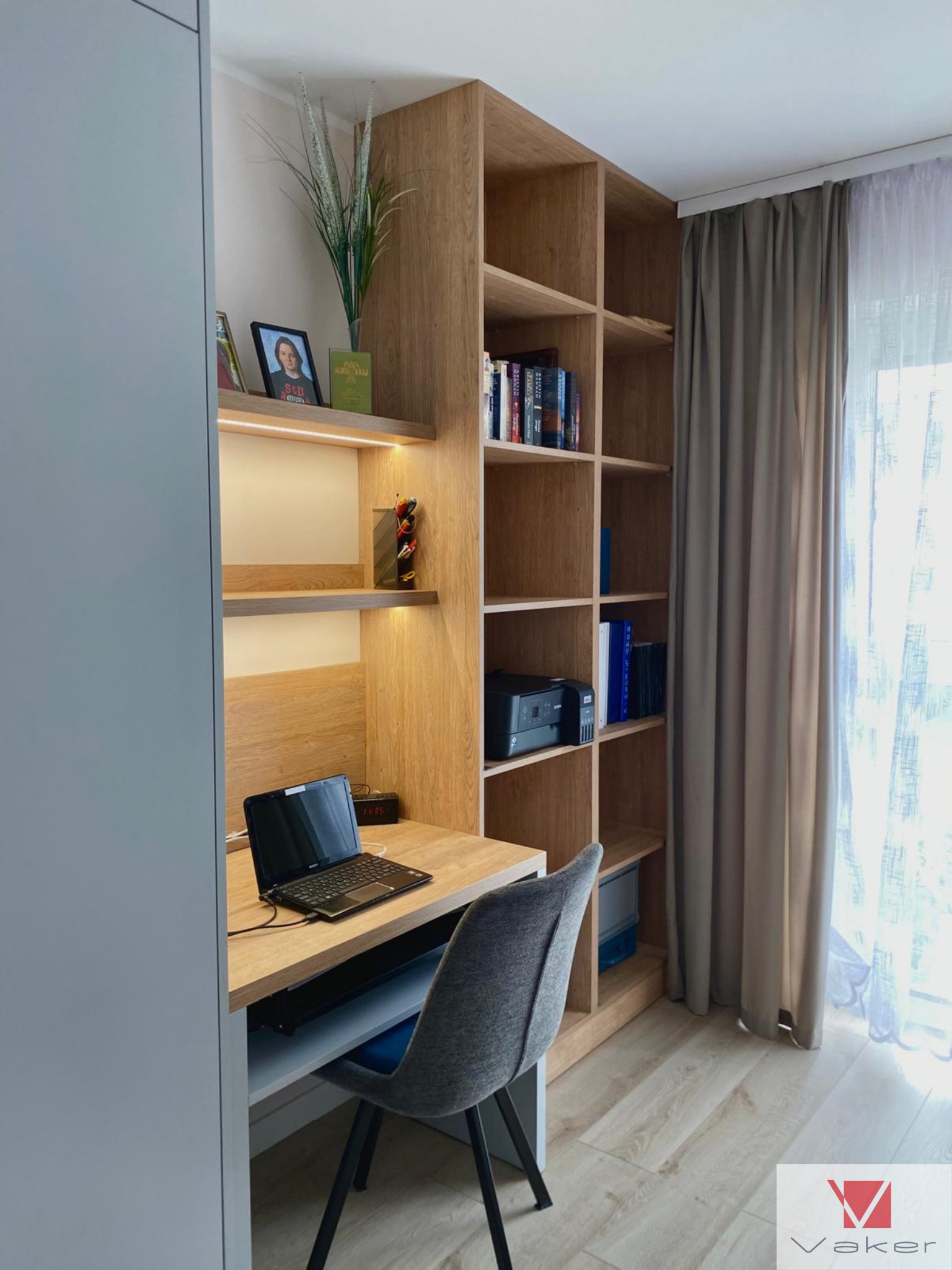 Drewniana zabudowa biurka z regałem i półkami podświetlanymi LED, szara tapicerowana krzesło, laptop na blacie, widok na okno z zasłoną.