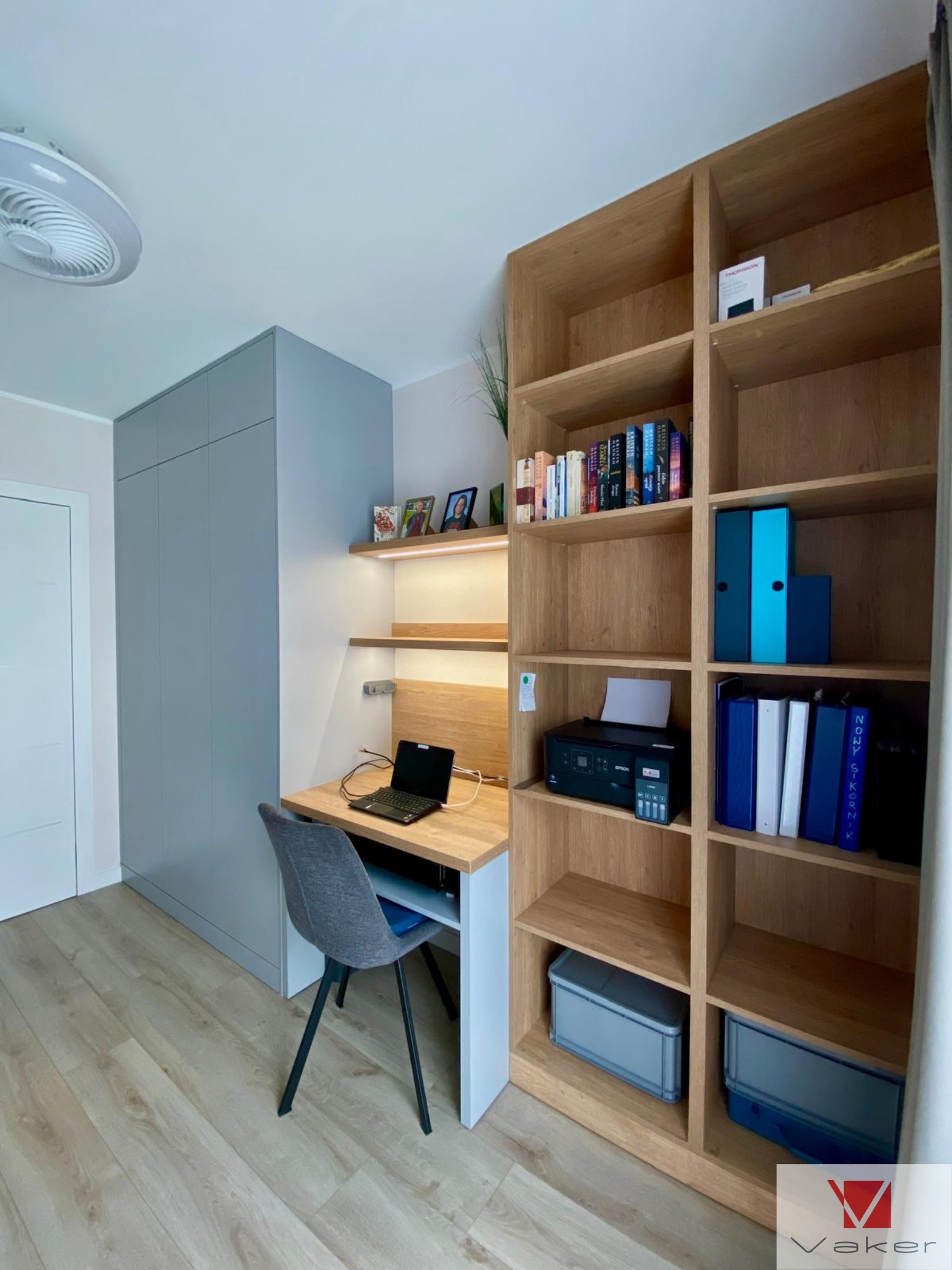 Pokój z szarą szafą na wymiar, biurkiem z oświetleniem i regałem z jasnego drewna z książkami i segregatorami.