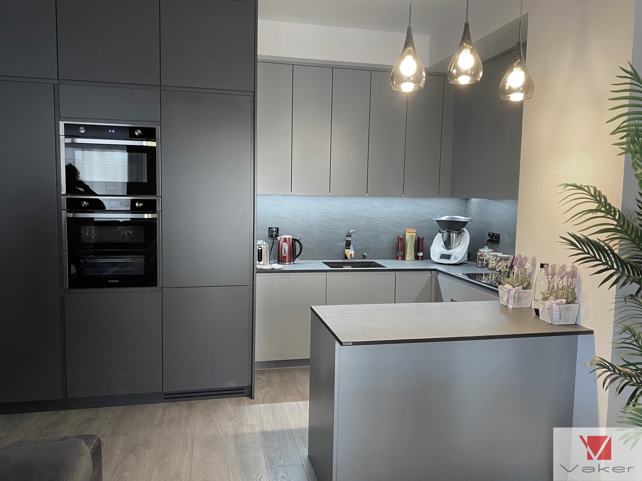 Nowoczesna kuchnia w odcieniach szarości z wyspą, oświetleniem punktowym i zabudowaną piekarnikiem oraz mikrofalówką Samsung, uzupełniona dekoracjami roślinnymi i logo firmy Vaker w prawym dolnym...