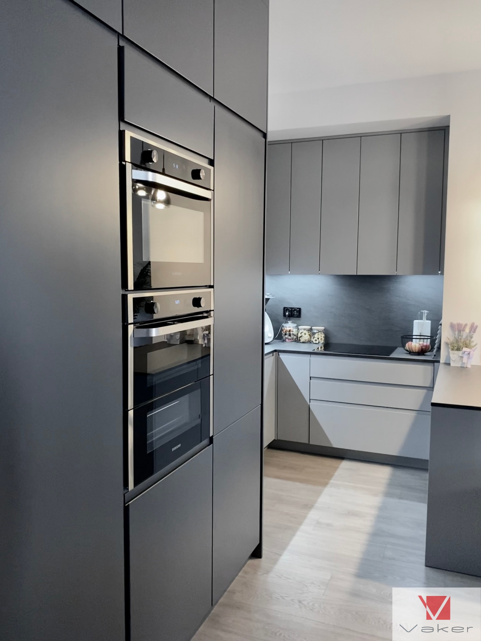 Nowoczesna szara kuchnia z zabudowanymi sprzętami AGD: piekarnik, mikrofalówka i płyta grzewcza. Minimalistyczny design z gładkimi frontami szafek i drewnianą podłogą.