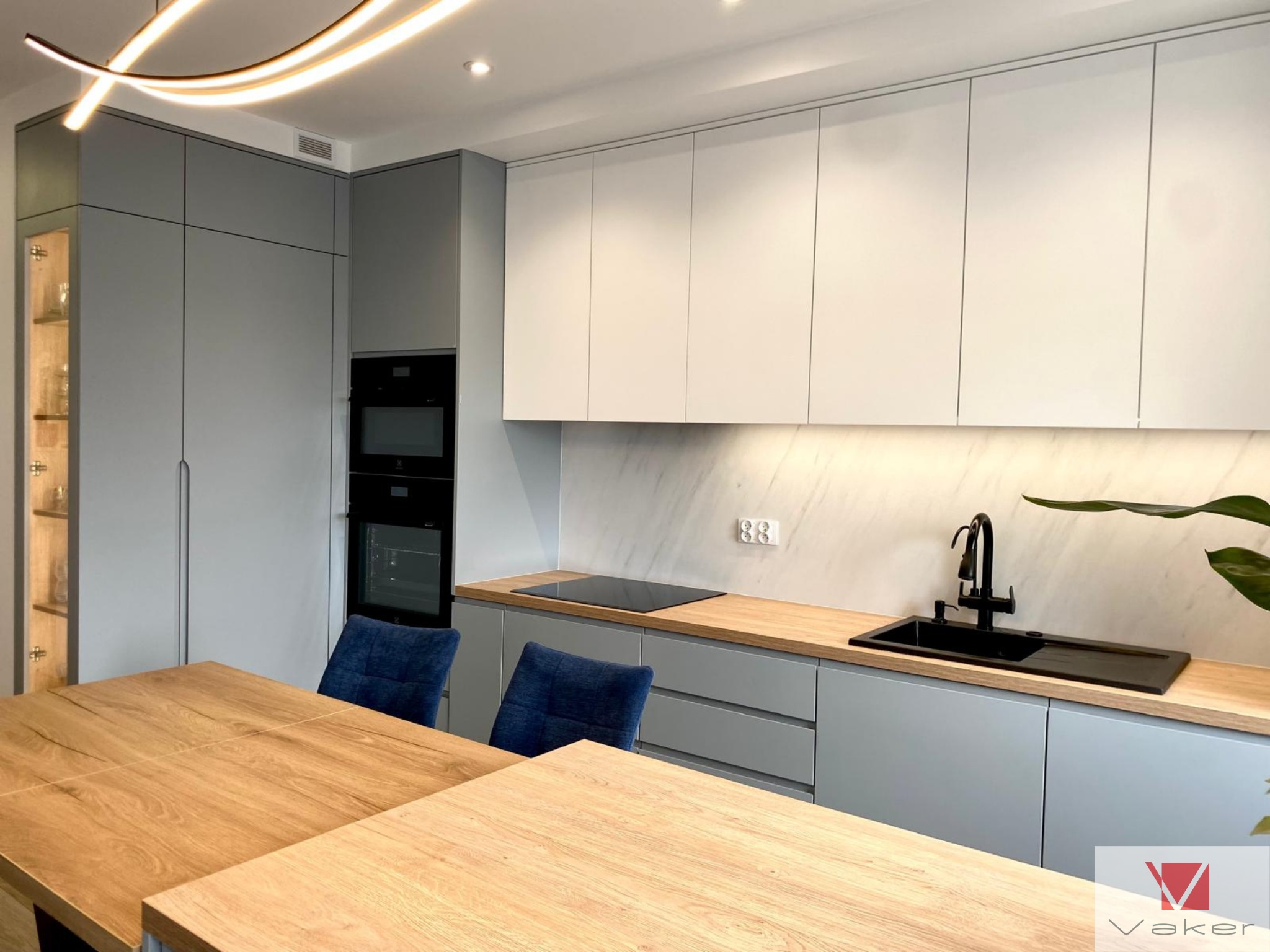 Nowoczesna kuchnia w odcieniach szarości i bieli, z drewnianym blatem, czarną armaturą i oświetleniem LED, widok na stół i krzesła.