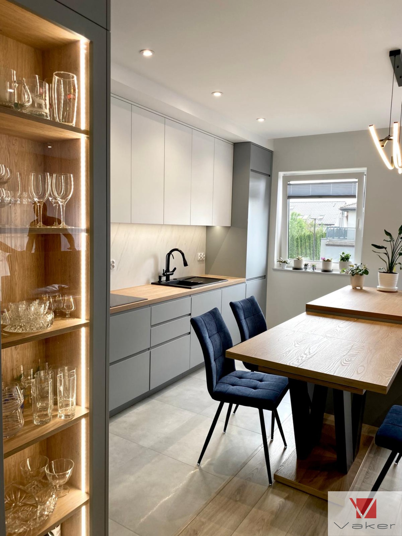 Nowoczesna kuchnia z szarymi szafkami dolnymi i białymi górnymi, drewnianym blatem, czarną armaturą, przeszkloną witryną z podświetleniem LED oraz drewnianym stołem z aksamitnymi krzesłami...